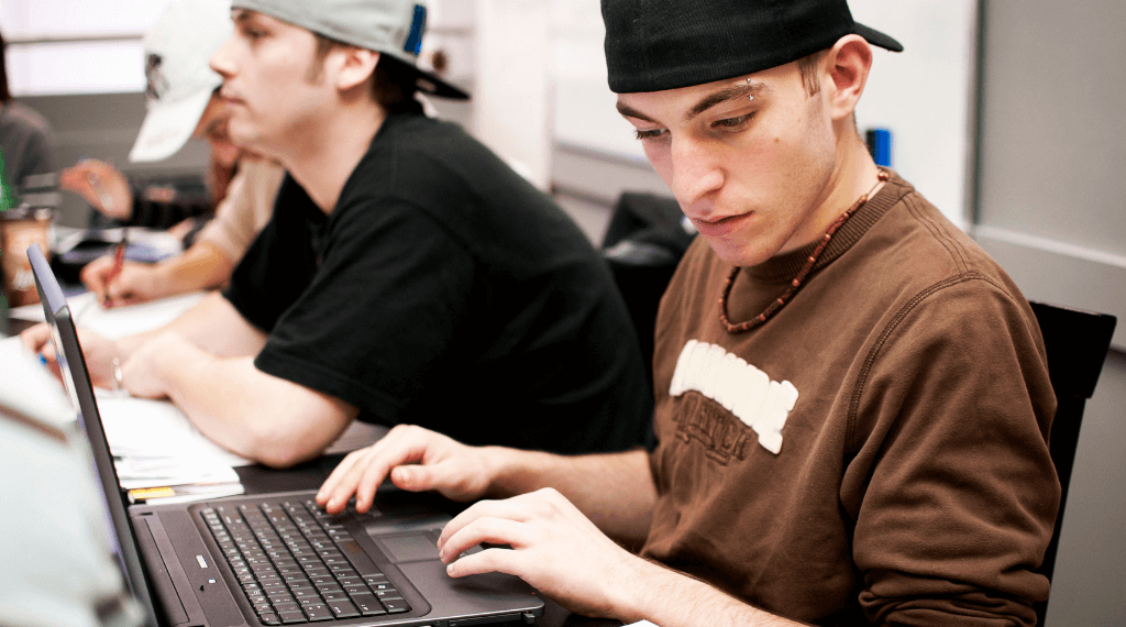 young adult man with hat backwards and brown shirt typing on laptop